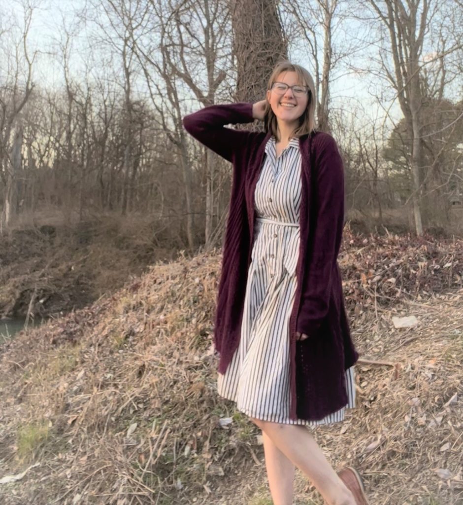 Woman in a stripped dress and maroon cardigan standing with trees all about her. 