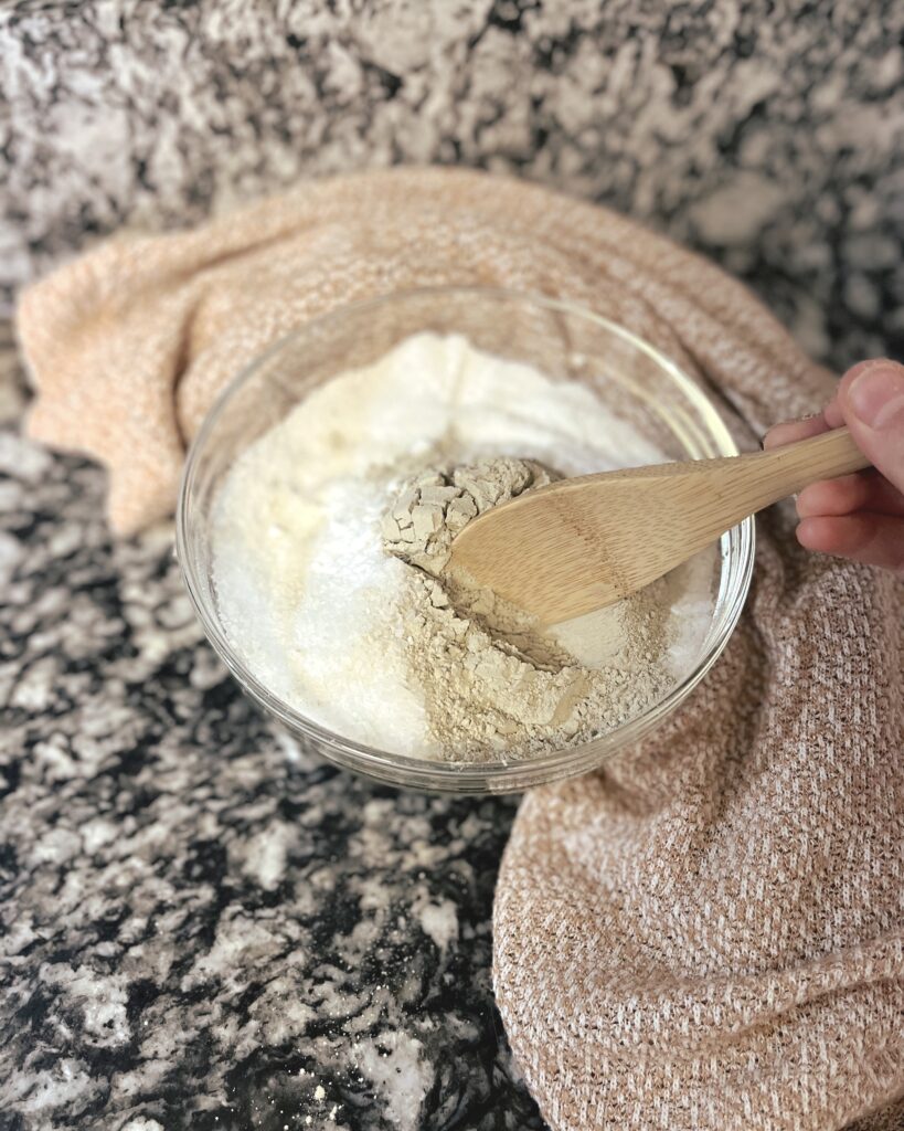 White Epsom salts and baking soda being mixed with grey clay power with a wooden spoon. 