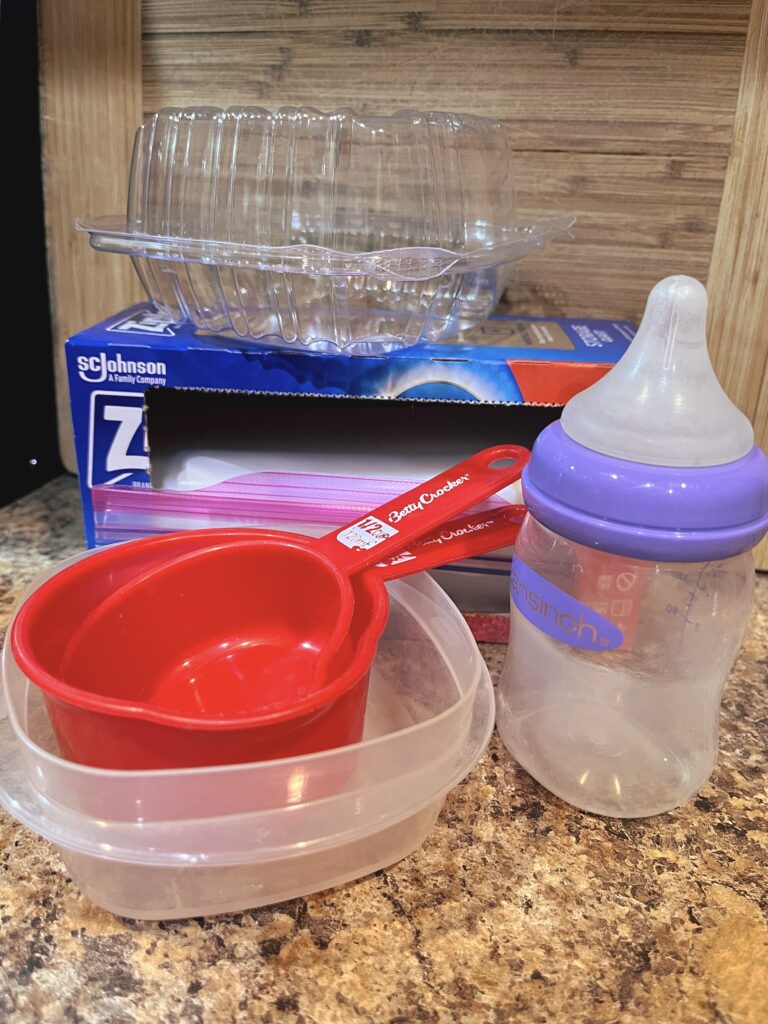 A plastic baby bottle next to two red measuring cups, a Tupperware container, and a box of plastic ziplock bags. 