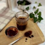 A jar full of homemade chocolate syrup on a cutting board next to a checked towel, the jar lid, and a chocolate covered spoon