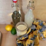 A swing top bottle full of water and another full of coconut water. A jar of milk, a lemon, and a lime sit in front of the bottles.