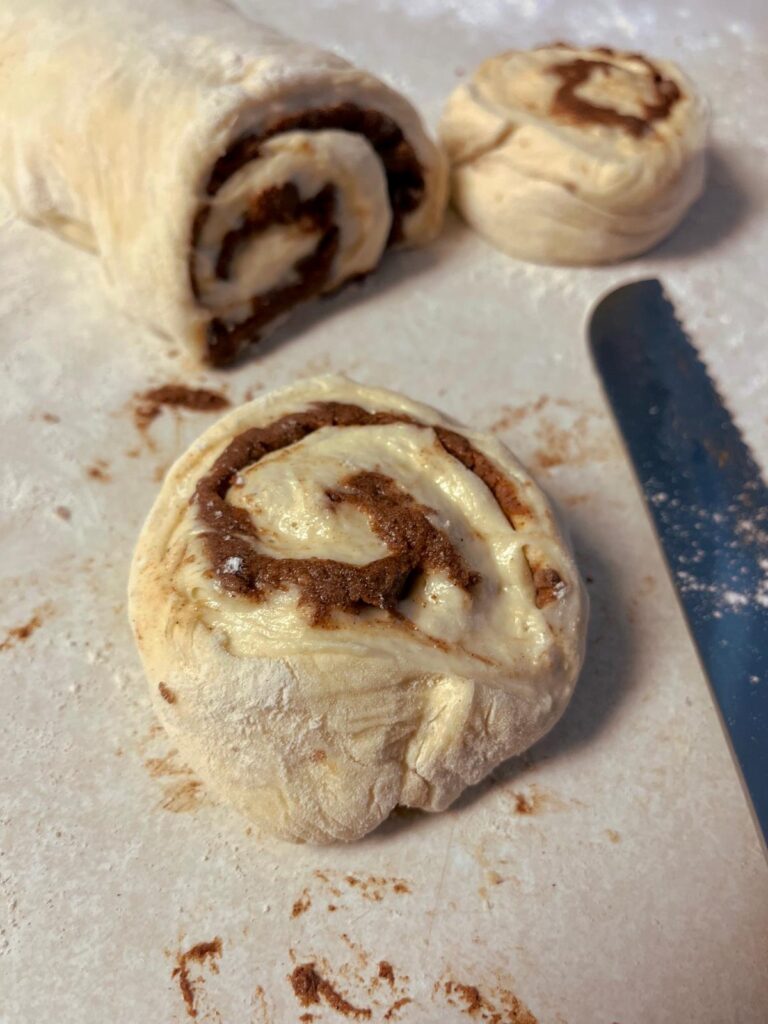 A cinnamon roll next to a log of dough and a serrated knife.