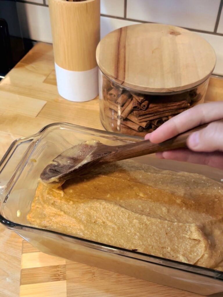 A wooden spoon being used to spread sourdough pumpkin bread into a glass loaf pan.