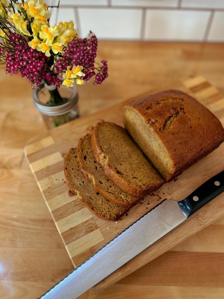 A sourdough pumpkin sweetbread on a wooden cutting board with a knife. A jar of wildflowers in the background