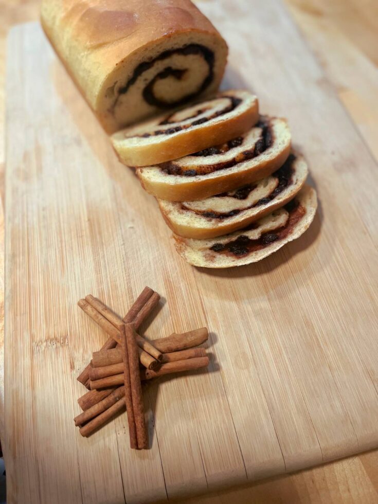 Sliced cinnamon raisin bread on a wooden cutting board next to cinnamon sticks.