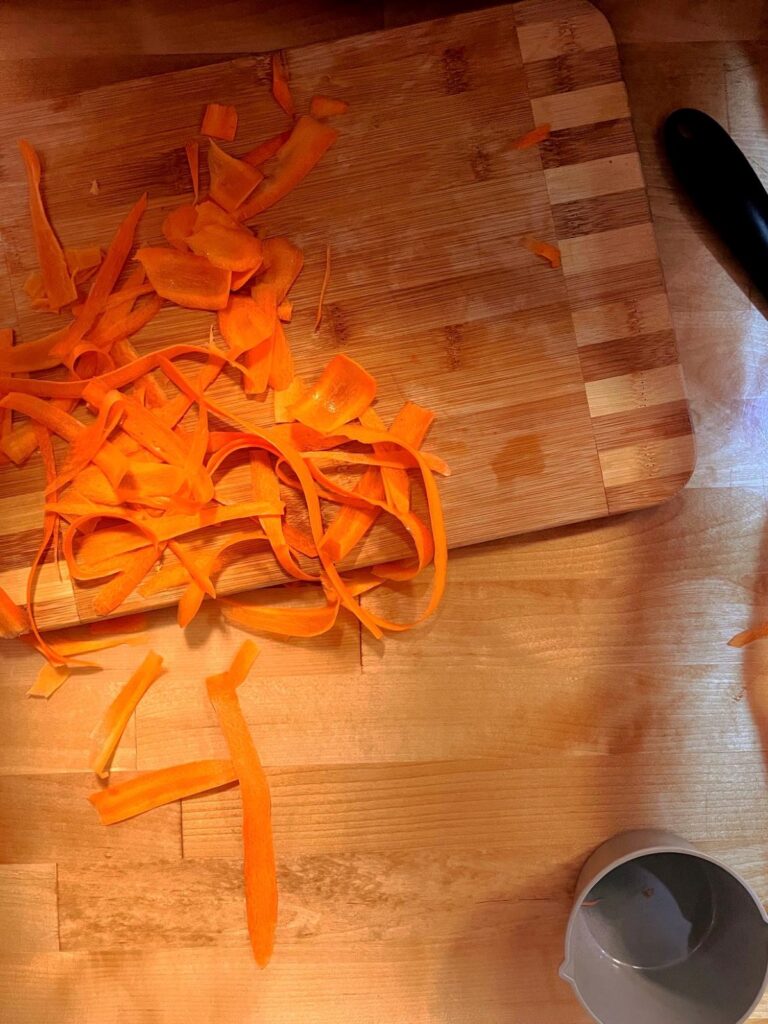 A bunch of ribboned carrots being prepped for a raw carrot salad laying on a wooden cutting board.
