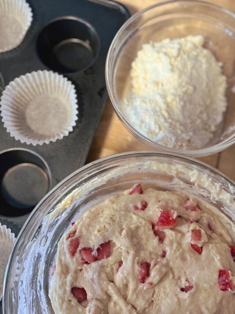 A bowl full of strawberry vanilla sourdough muffins next to a muffin tin and a bowl of strudel topping.