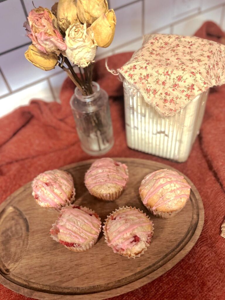 Strawberry vanilla sourdough muffins on a wooden cutting board next to sourdough starter and dried roses.