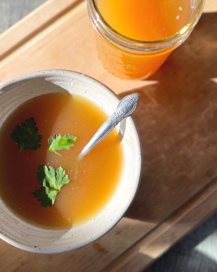 A ceramic bowl and glass jar filled with homemade chicken bone broth on a cutting board in the sun.