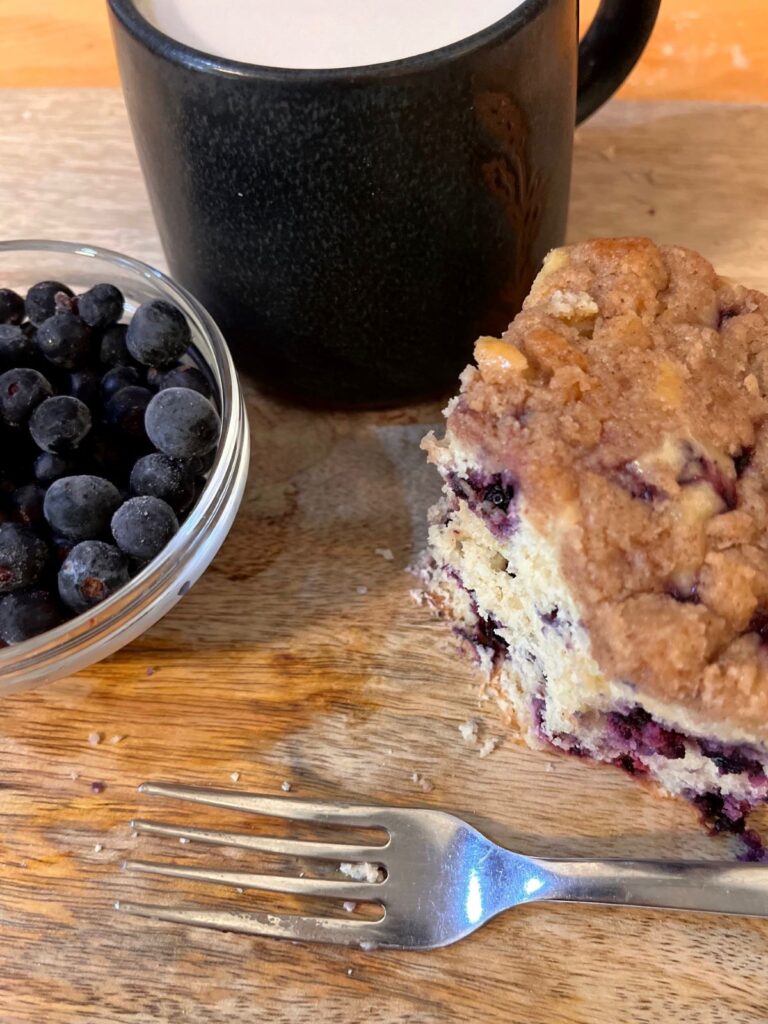 A slice of sourdough blueberry buckle with a bowl of fresh blueberries, a fork, and a black mug of tea, on a black cutting board.