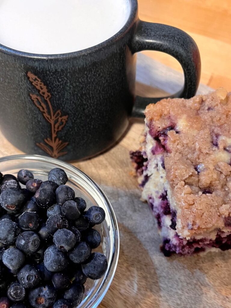 A bowl of blueberries, a slice of sourdough blueberry buckle, and a mug of tea on a wooden cutting board.