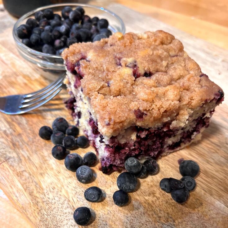 A square of blueberry coffeecake on a wooden cutting board with a bowl of blueberries and a fork.