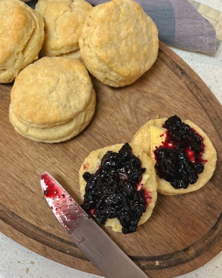 An oval wooden cutting board with a pile of sourdough biscuits, one is cut open with jam and butter