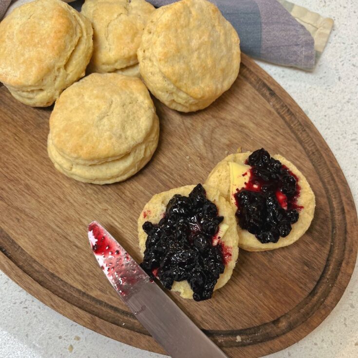 An oval wooden cutting board with a pile of sourdough biscuits, one is cut open with jam and butter