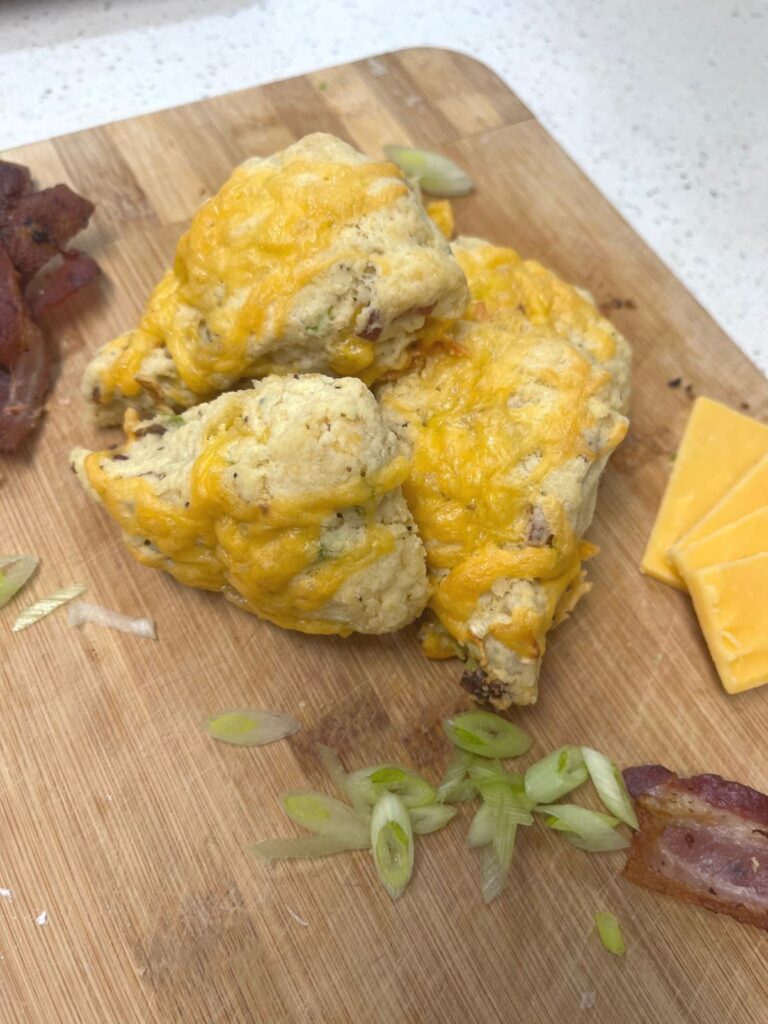 A pile of savory scones on a cutting board with bacon, cheddar cheese, and sliced green onion.