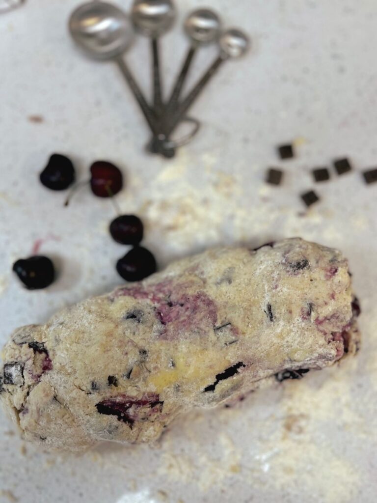 A lump of sourdough scone dough on a floured countertop with measuring spoons, cherries, and chocolate chunks.