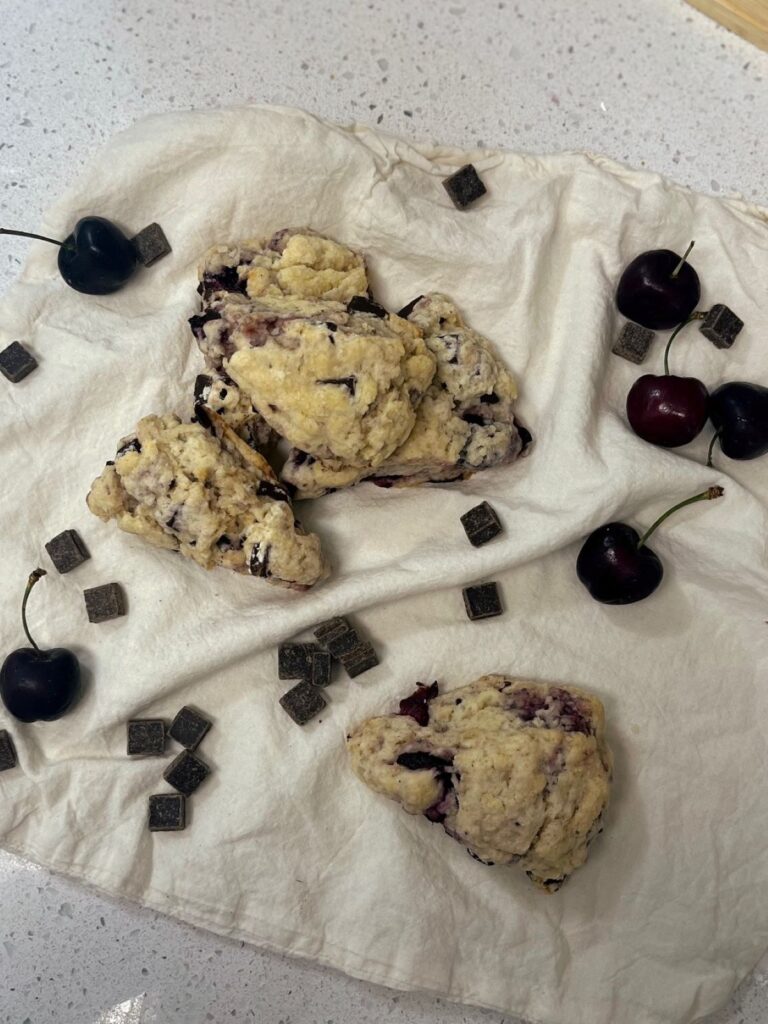 Three chocolate cherry sourdough scones on a white cloth with chocolate chunks and cherries all around.
