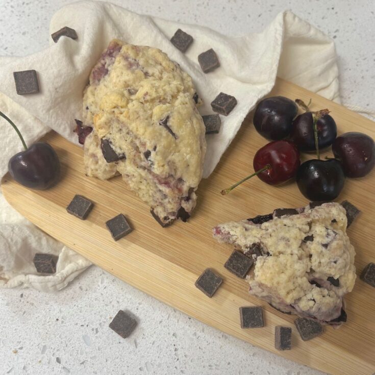 Two chocolate cherry sourdough scones on a cutting board with a pile of cherries and chocolate chunks scattered