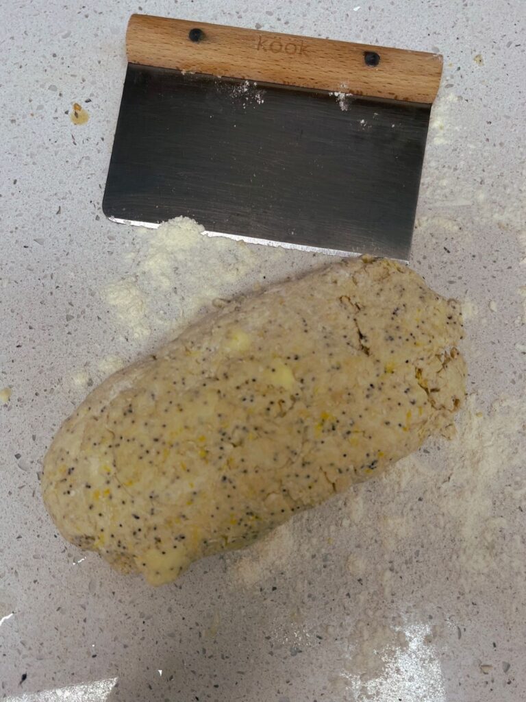 A log of lemon poppy seed scone dough with a bench scraper on a counter.