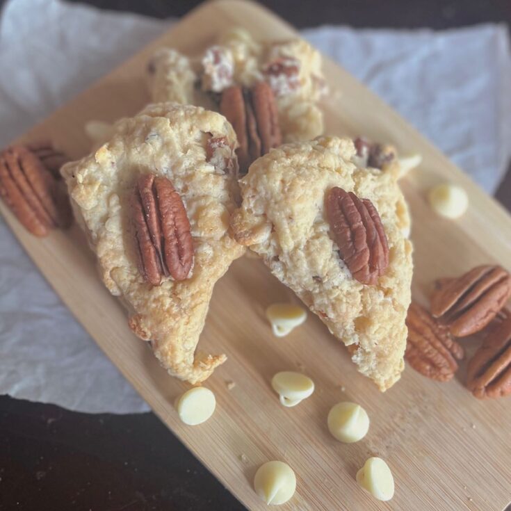 A pile of scones with a pecan on the center of each one on a wooden cutting board with pecans and white chocolate chips