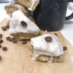 A pair of coffee sourdough scones on a piece of parchment paper with coffee beans scattered around and a black mug with coffee.