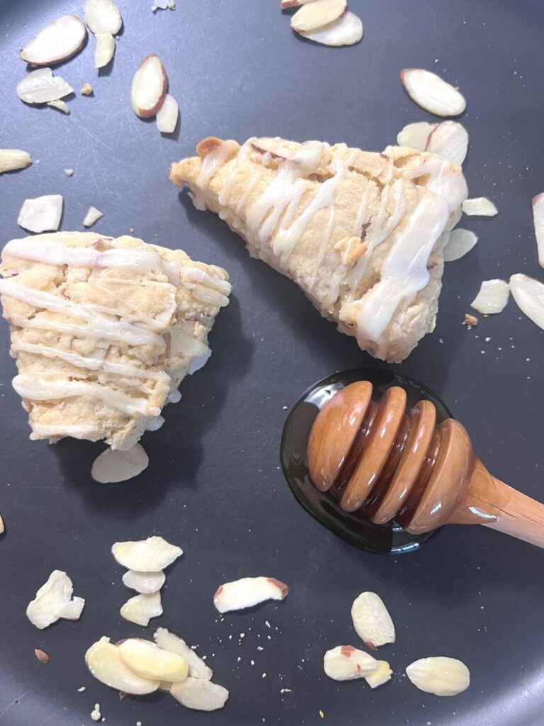 Two sourdough scones on a black plate with a wooden honey dipper and almonds scattered around.