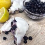 Two blueberry sourdough scones on a cutting board with a bowl of blueberries and some lemon halves.
