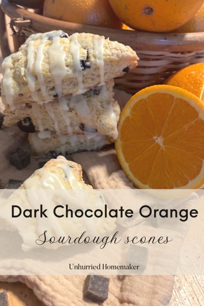 A stack of sourdough scones in front of a basket of oranges surrounded by cut oranges and chocolate chunks