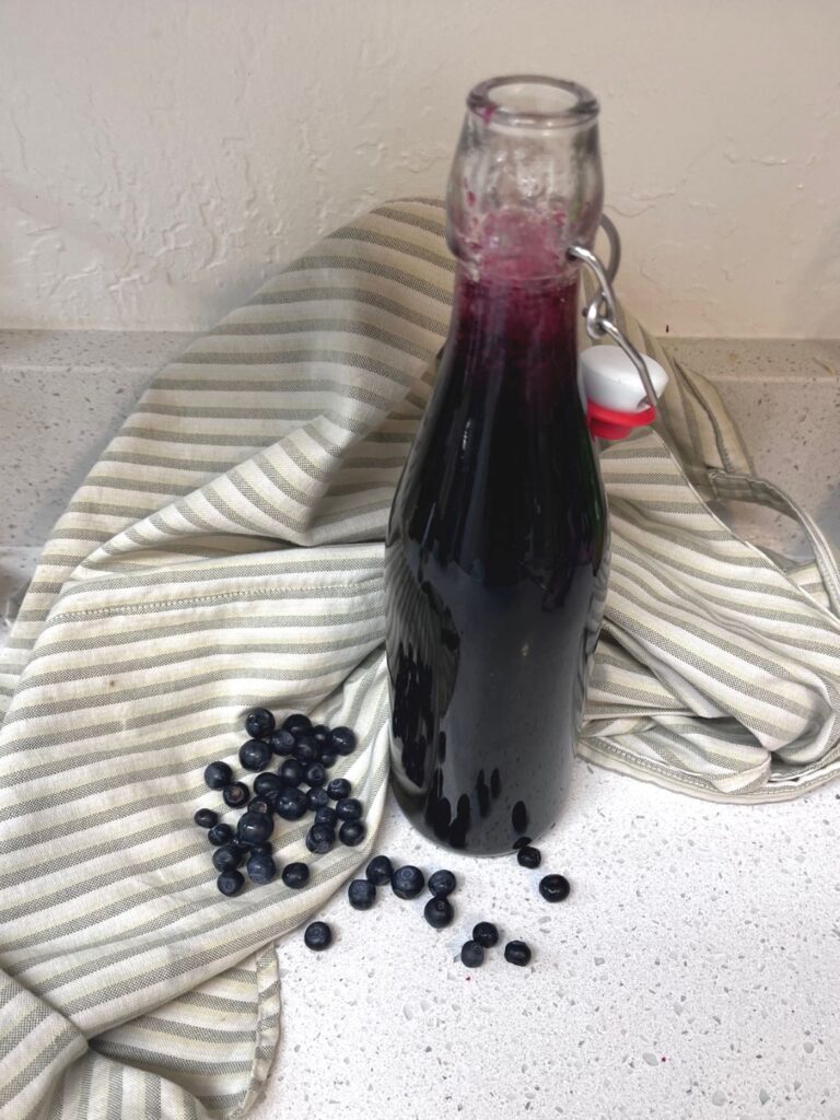 An open swing top bottle of blueberry syrup with a stripped cloth behind it and some blueberries scattered around.