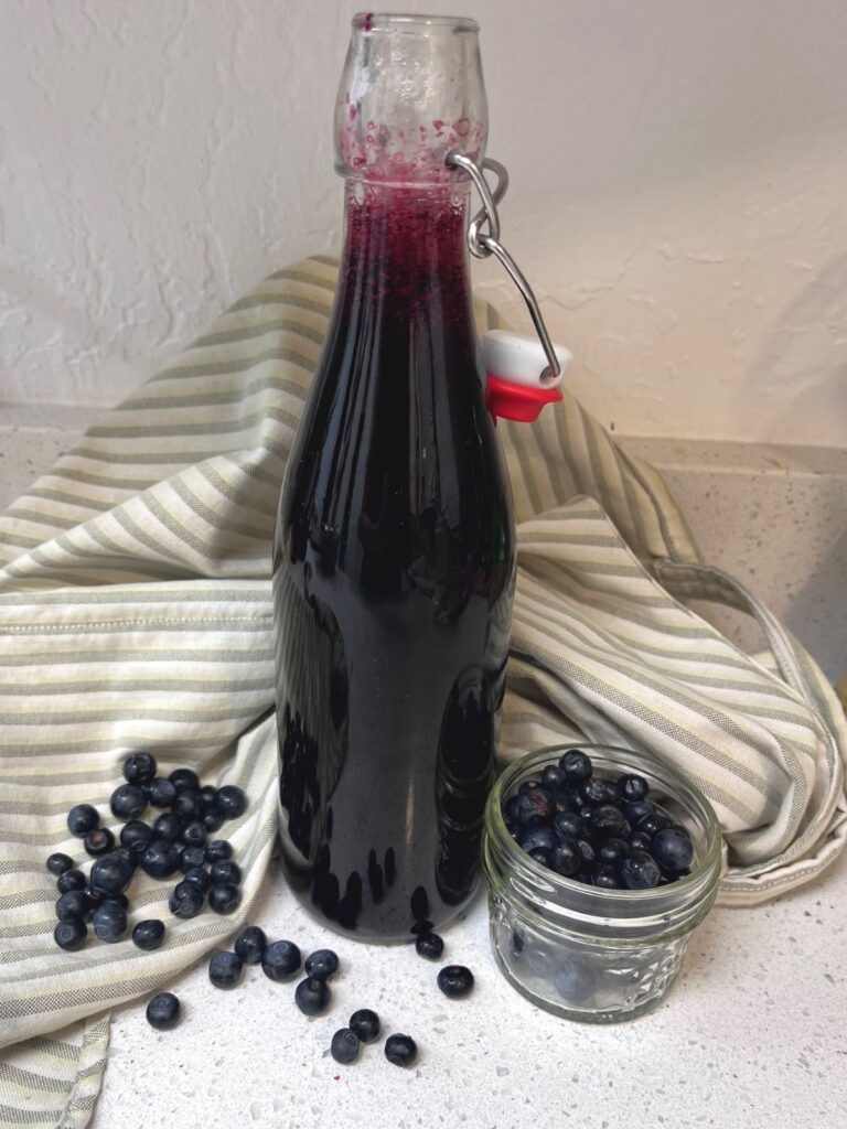 A tall bottle of blueberry simple syrup in front of a stripped cloth. A small jar of blueberries and some scattered around the bottle.