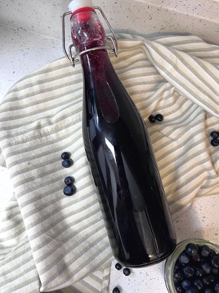 A bottle of blueberry simple syrup laying on a stripped cloth with a jar of blueberries and some scattered around.