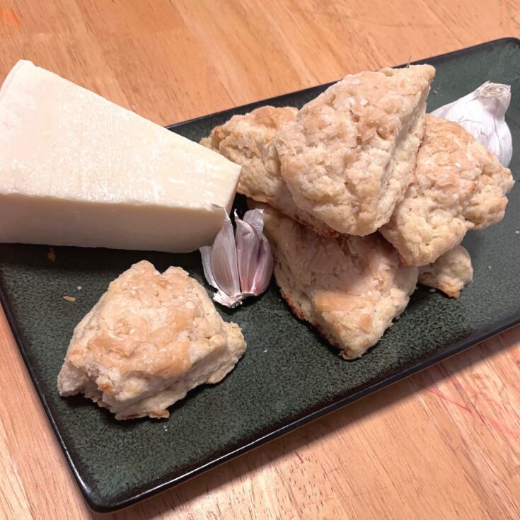 A green rectangle plate with a wedge of parmesan, some garlic, and a pile of garlic parmesan sourdough scones.