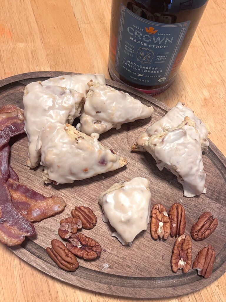 A dark wooden cutting board with scones, bacon,a nd pecans, and a bottle of maple syrup in the background.