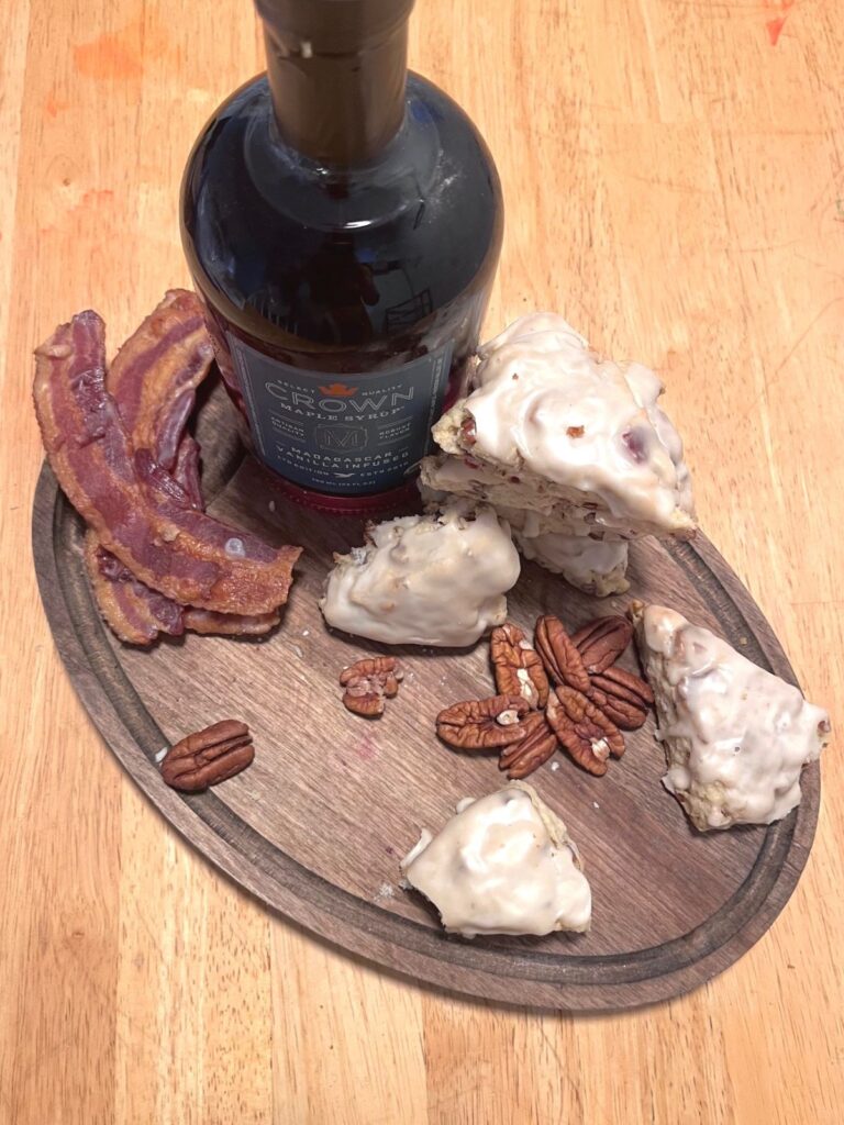 A bottle of maple syrup, a stack of glazed sourdough scones, strips of bacon, and pecans on a dark wooden board.
