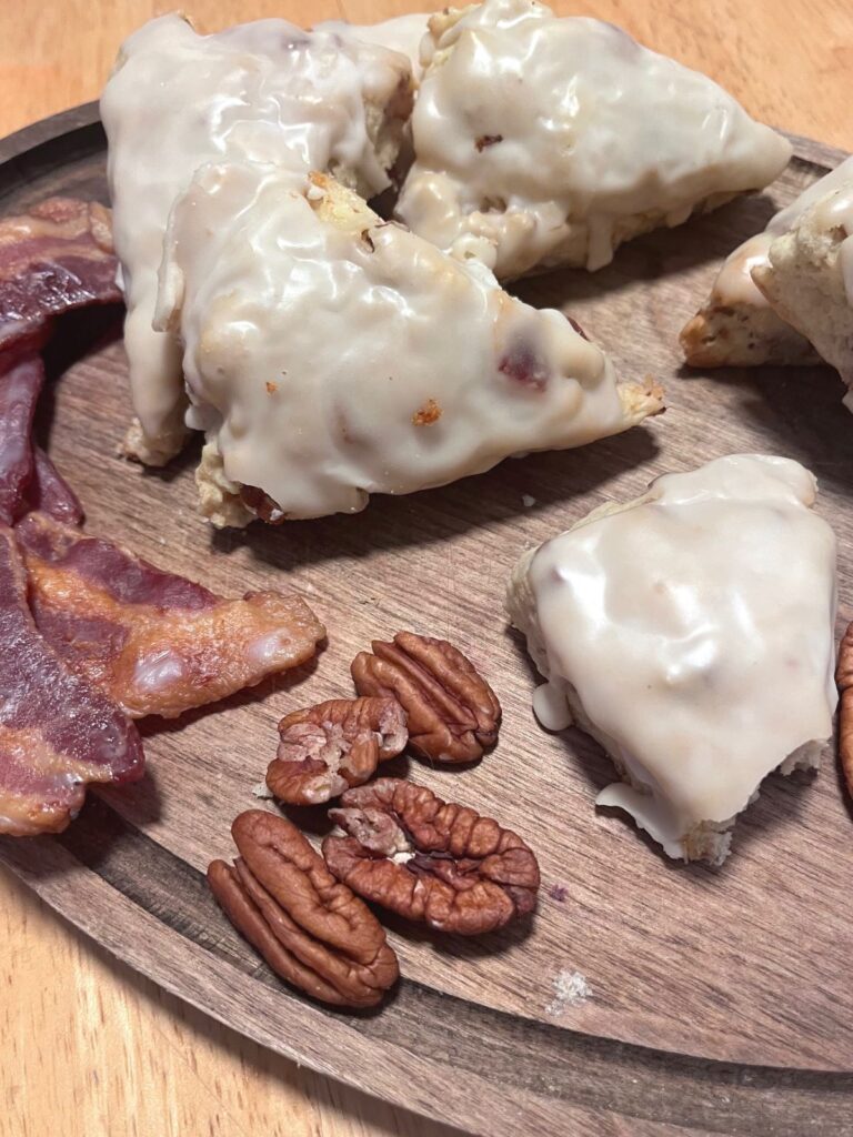 A pile of glazed scones with strips of bacon and a few pecans on a wooden cutting board