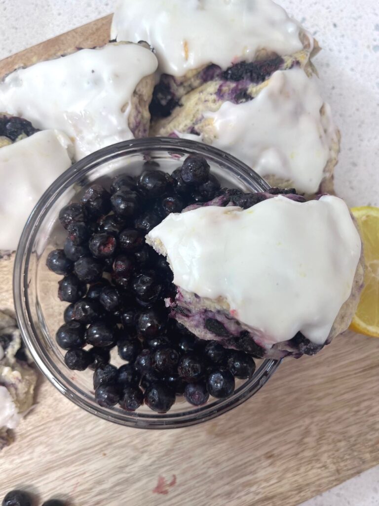 A scone balanced on the rim of a bowl of blueberries with other scones in the background.