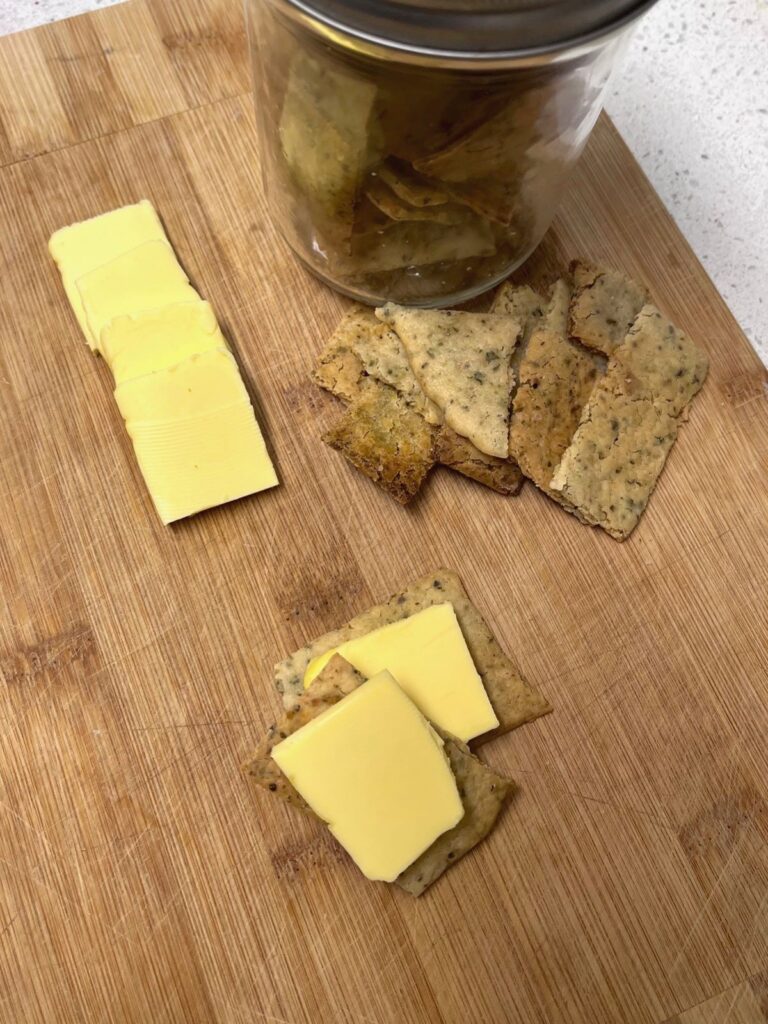 A pile of sourdough crackers next to a jar of crackers and a few slices of cheese.