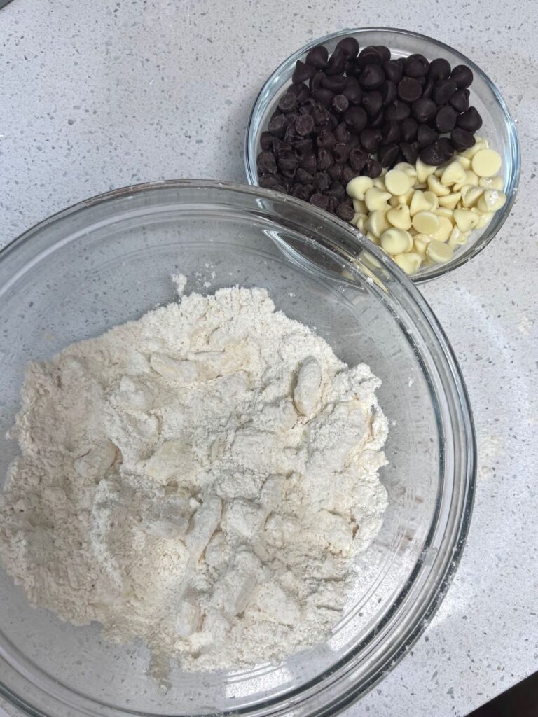 A glass bowl full of dry ingredients and another bowl with three types of chocolate chips