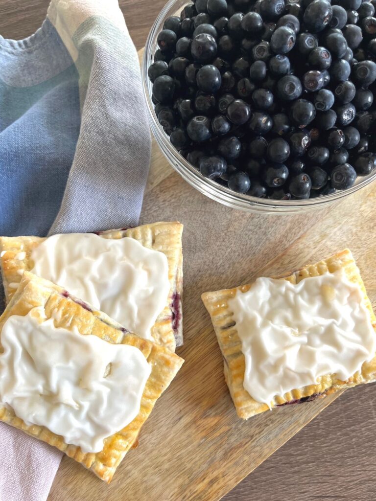 Thre blueberry poptarts on a cutting board with a bowl of fresh blueberries.