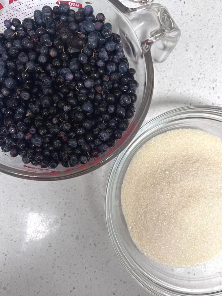 A large liquid measuring cup of blueberries next to a glass bowl of sugar.