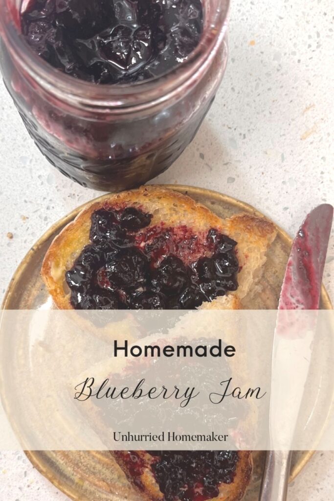 A slice of toast with homemade blueberry jam on it next to a jar of jam and a butter knife. 