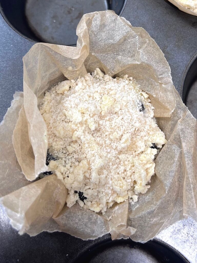 An unbaked muffin in a muffin tin with crumb topping over the top of the muffin.
