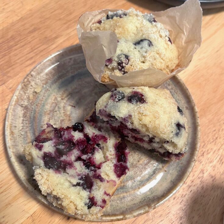 A bakery style, sourdough blueberry muffin on a plate nest to another muffin cut in half.