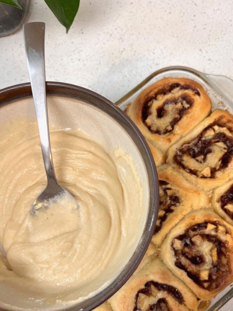 A bowl of glaze over a pan of freshly baked apple cinnamon rolls.