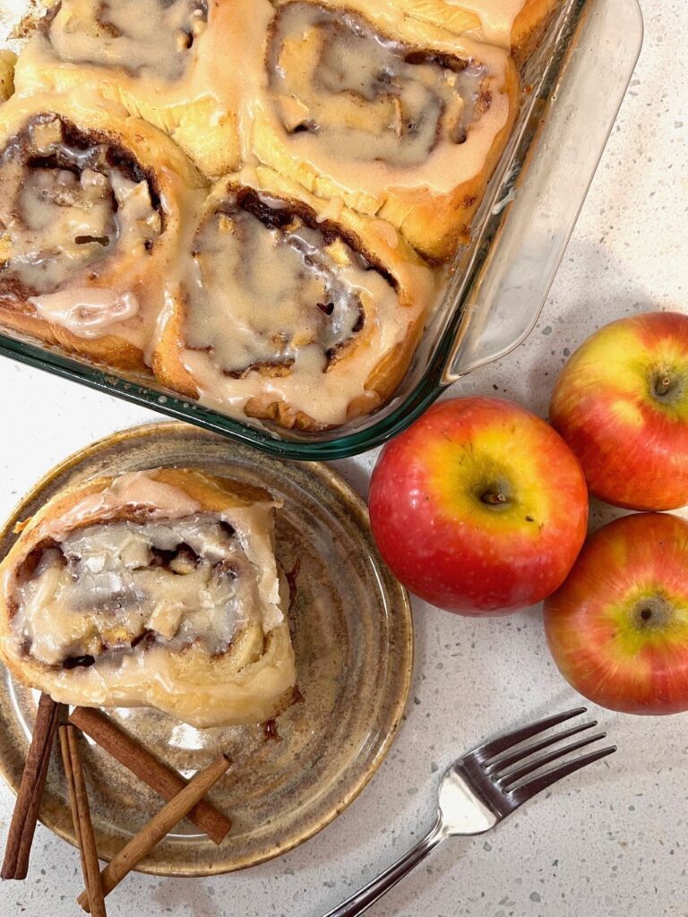 A pan of cinnamon rolls, with one on a plate with cinnamon sticks, and three red apples on the counter next to them.