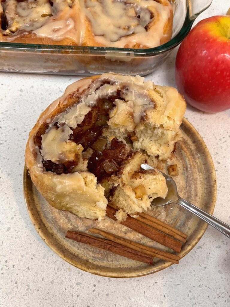 A soft cinnamon roll being broken open with a fok on a plate with cinnamon sticks and apples in the background.