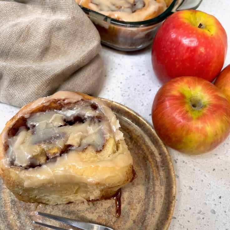 A sourdough apple cinnamon roll on a plate with the pan behind it and three red apples next to the plate.