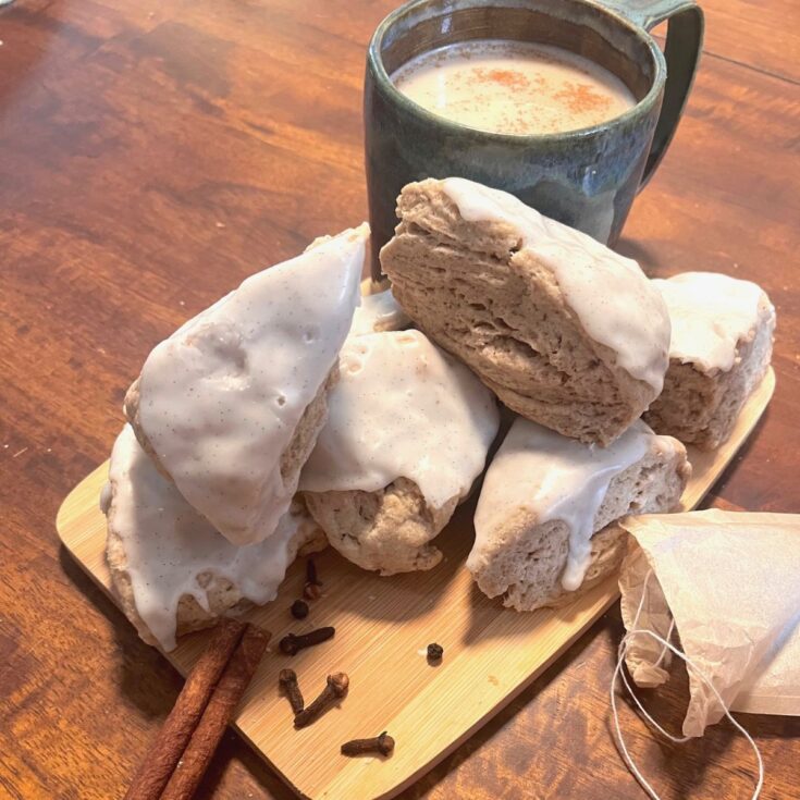 A small wooden cutting board piled with whole spices, sourdough chai scones, and a ceramic mug of chai tea.