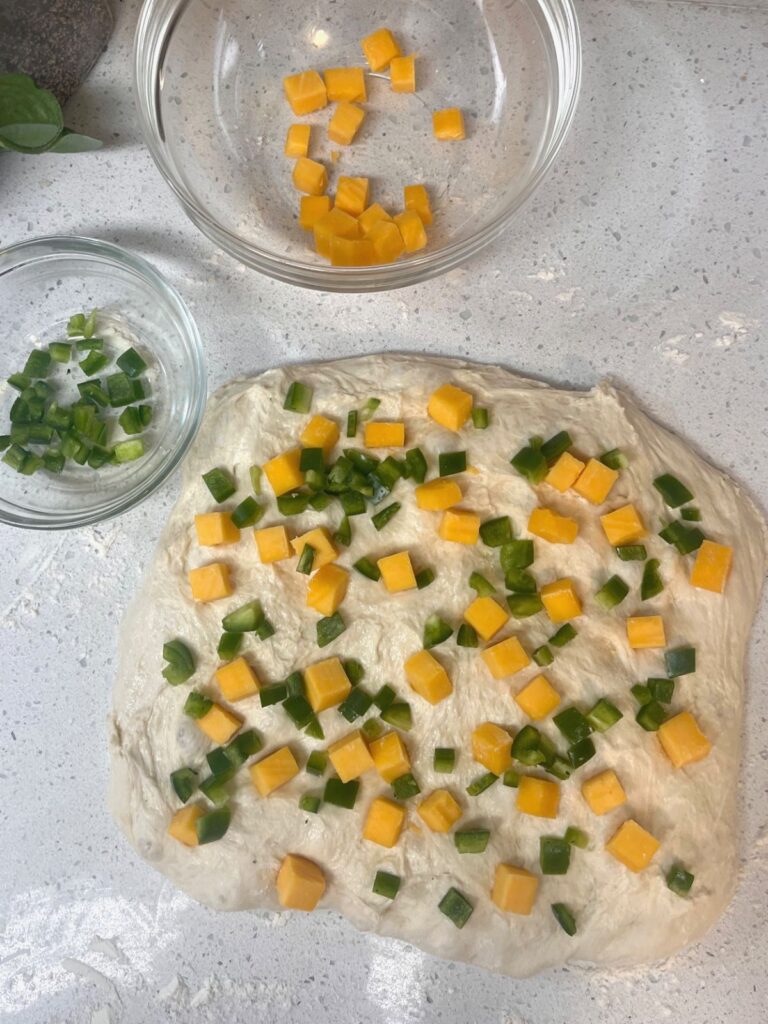 A rectangle of bread dough with cheddar cheese and jalapenos spread on it.