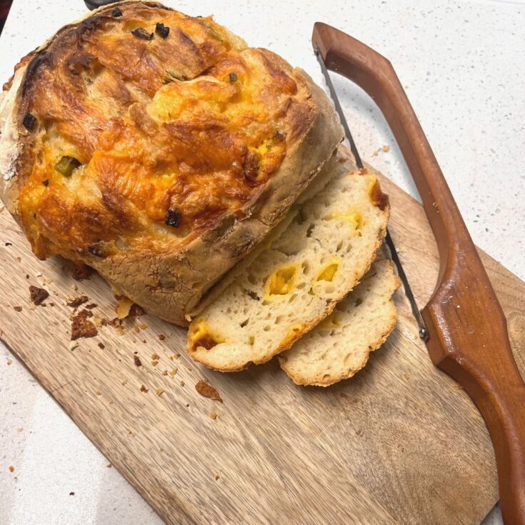 A loaf of cheddar jalapeno sourdough bread on a wooden cutting board with a bow knife.